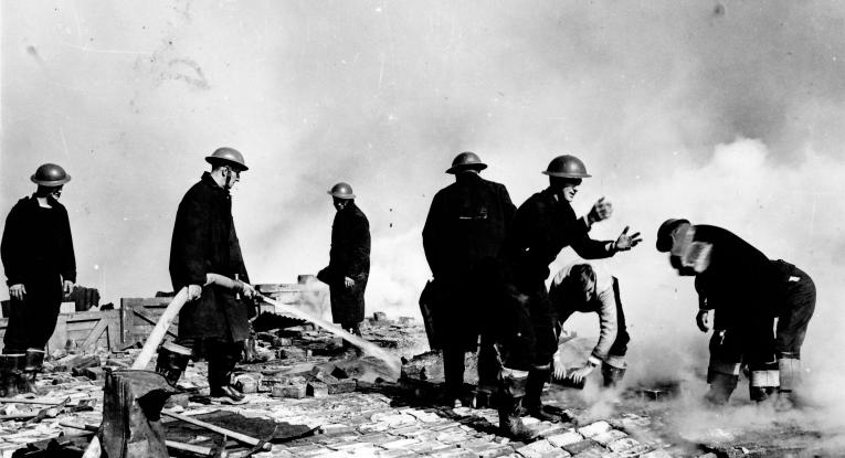 What looks to be eight firefighters in the streets, which are ablaze. They are in full uniform (jumpsuits and helmets). One is holding a hose, one is reaching to catch something, two are stood looking around and others are crouched down handling bricks.