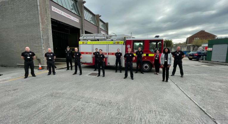 Twelve members of the Service stood in front of a fire appliance on a grey cloudy day. The six in the middle are the on-call apprentices, in their black uniform. Other firefighters are stood to the left and right, as well as two female support staff.