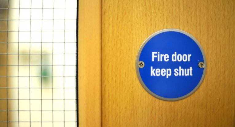 The blue circular sticker in a fire door which reads 'fire door keep shut' on a wooden fire door, with a but of the grid window of the fire door in the photo.