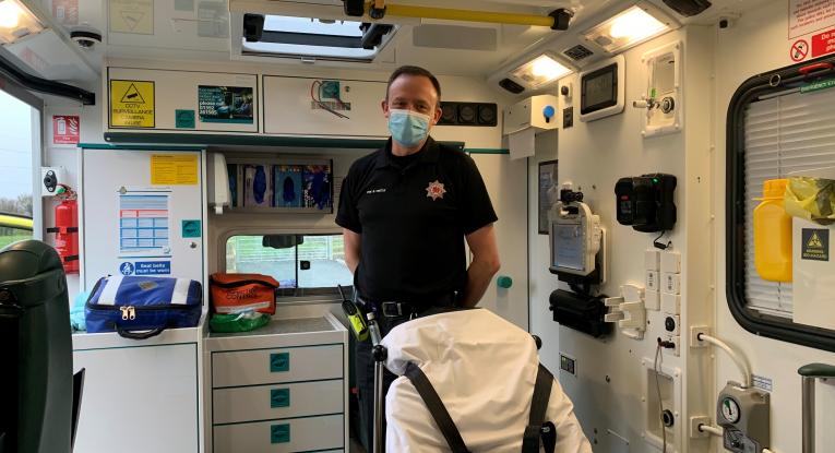 One of our firefighters, Robert Partridge, stood in the back of an ambulance surrounded by lots of ambulance equipment. He has his firefighter uniform on (which is a black shirt and trousers) and a mask.