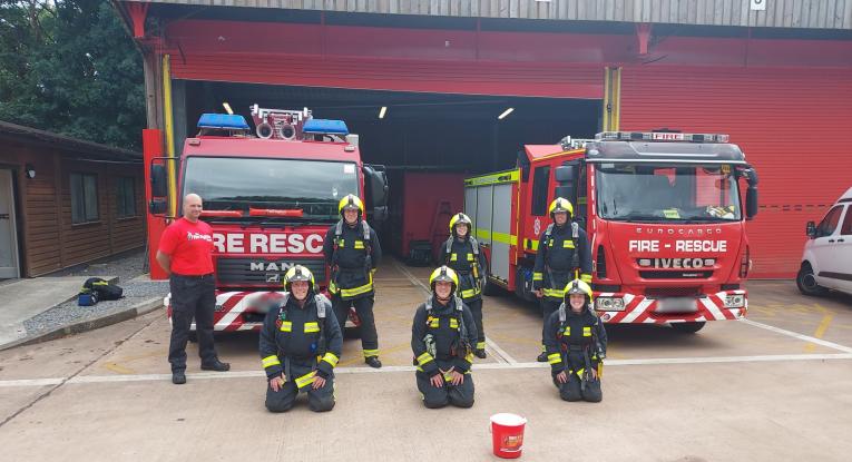 Seven people stood in front of two fire engines with an open station red garage behind them. Three are knelt on the ground, and four are stood behind. One is in a red Fire Fighters charity shirt whilst the others are in full kit. 