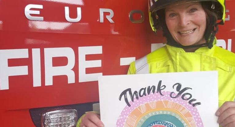Sally stood in front of a fire engine in her firefighter kit, which is a neon jacket and matching helmet. She's smiling and holding an A4 piece of paper which has 'thank you' written on it alongside a rainbow with purple, orange, green, blue and red.