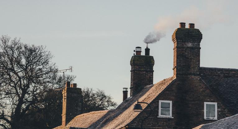 A few chimneys in a row, with smoke coming out of the one in the middle. Only the roofs of the houses can be seen, as well as some of the brick wall of the first in the row, and a grey sky. There are a few trees in the background with no leaves, so it's clearly autumn.