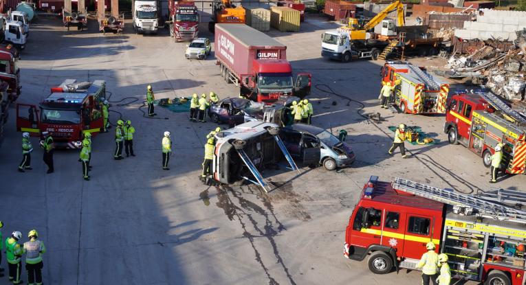 A concrete space being used for road safety training. There are lorries around the edge, four fire engines, firefighters dotted around and a model of a road traffic collision in the middle between three cars, a van and a lorry. A police car is also nearby.