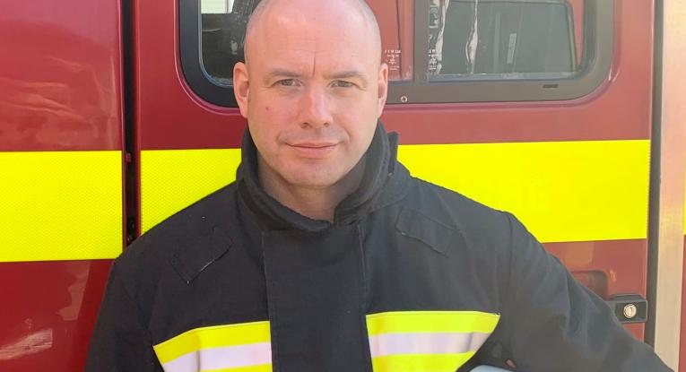 A male firefighter stands in front of a fire engine in uniform. 