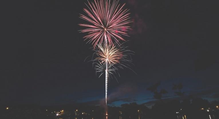 A red firework in the night sky, with orange and yellow smaller fireworks directly beneath it. The display looks like it's happening near a body of water.