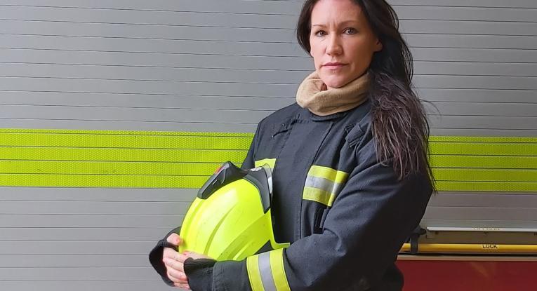 A close-up of firefighter Laura stood in her kit holding her helmet.