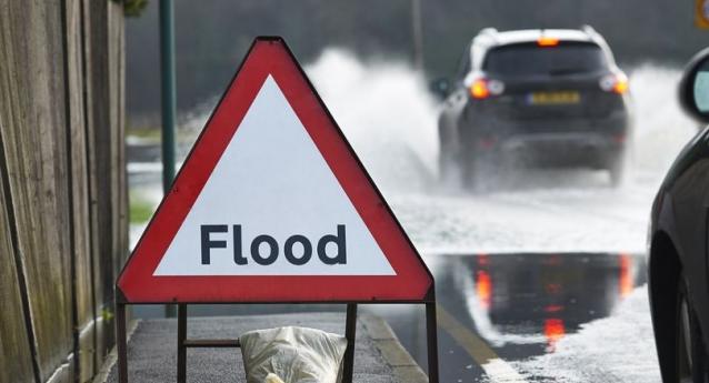 A flood warning sign displayed on next to a main road with cars driving through floodwater. 